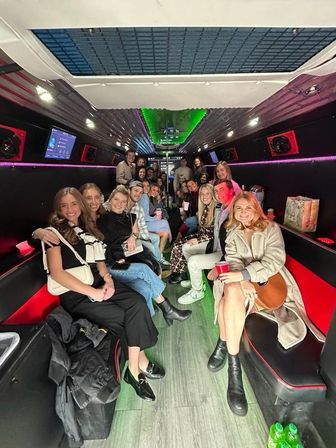 Large group of friends smiling and holding cups inside a neon-lit party bus with leather bench seats, LED ceiling lights and wood-look floor — lively night-out celebration scene.