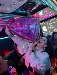 Laughing woman in a fringe jacket holding an oversized pink "Bachelorette" balloon inside a neon-lit party bus with friends, disco lights, and a karaoke screen