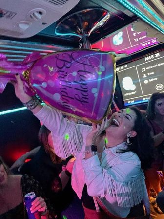 Laughing woman holding a giant purple bachelorette champagne-bottle balloon inside a neon-lit party bus with friends and a karaoke screen