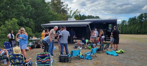 Outdoor tailgate-style gathering around a black motorhome in a grassy gravel clearing, people chatting and snacking under an extended awning with folding chairs, coolers and picnic gear, tree line and cloudy sky in the background.