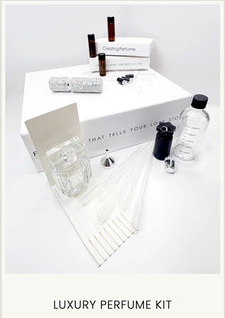Flatlay of a luxury DIY perfume kit on white background: white gift box, glass perfume bottle, amber sample vials, clear measuring bottle, pipettes, funnel and a glittery roll — fragrance-making tools
