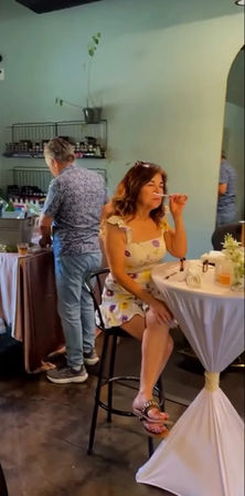 Woman in a yellow floral dress sits on a barstool, sniffing a fragrance blotter at a draped cocktail table during a casual indoor tasting or boutique event, with shelves of jars and a man browsing in the background.