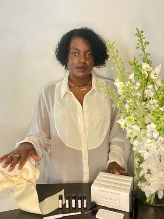 Woman with short curly hair wearing a sheer white blouse stands indoors behind a table of perfume samples, boxed fragrances with a cream ribbon and a tall white floral arrangement.