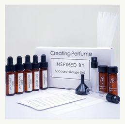 Fun DIY perfume-making kit on white background: white 'Creating Perfume' box with amber dropper vials labeled scent notes (spicy citrus, light floral, amberwood, leather, sweet moss), two small sample bottles, funnel, spray atomizer, scent strips and a blending worksheet.
