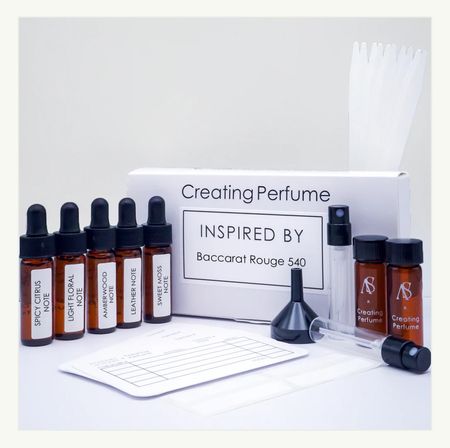 Fun DIY perfume-making kit on white background: white 'Creating Perfume' box with amber dropper vials labeled scent notes (spicy citrus, light floral, amberwood, leather, sweet moss), two small sample bottles, funnel, spray atomizer, scent strips and a blending worksheet.