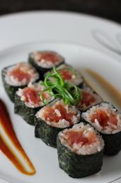 Close-up of fresh tuna maki sushi rolls wrapped in nori with sticky rice, topped with curled green onion garnish and brown and reddish sauce drizzles on a white plate