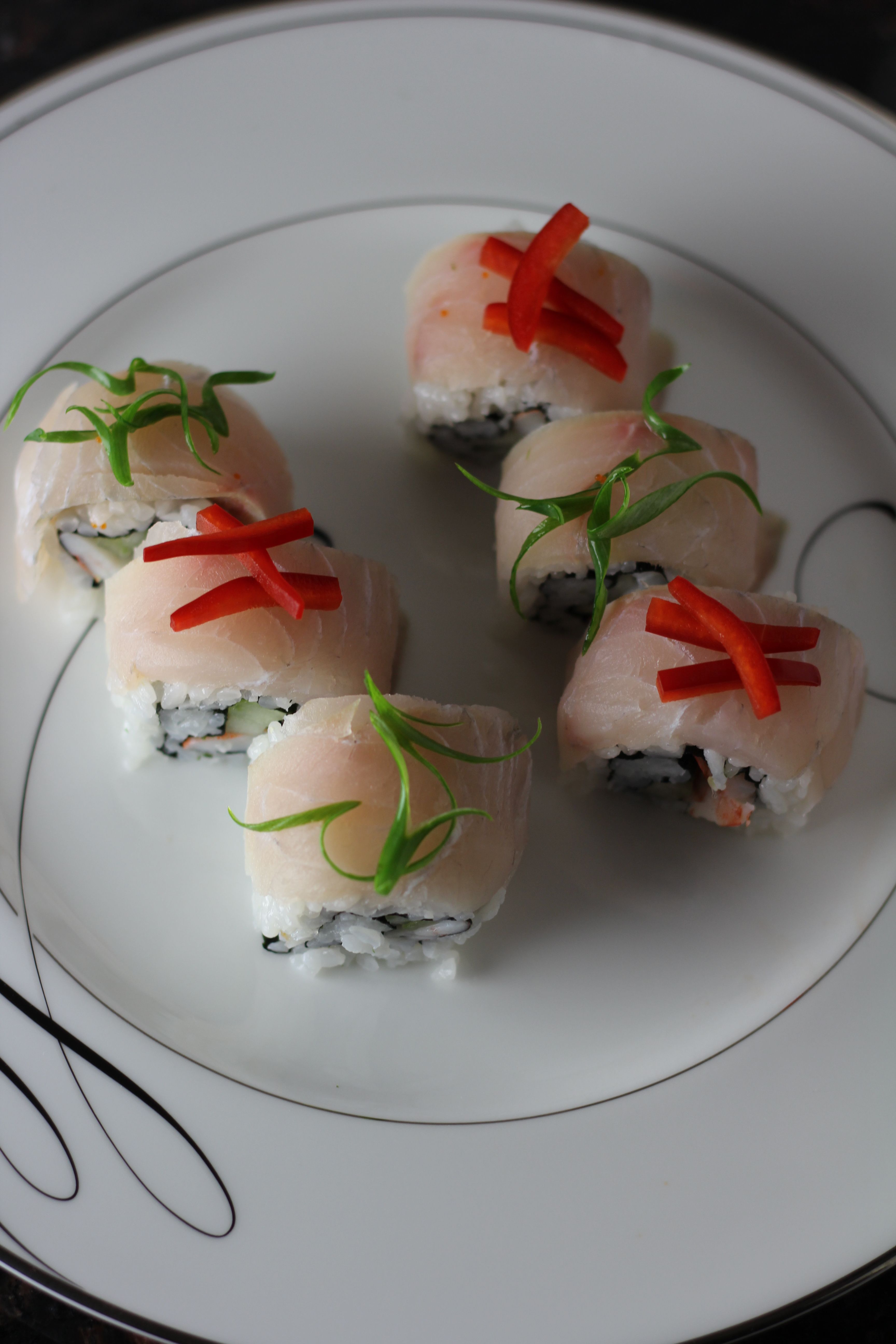 Close-up of six gourmet sushi rolls on a white porcelain plate, topped with thin white fish, red pepper strips and delicate green scallion curls