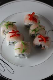 Close-up of six gourmet sushi rolls on a white porcelain plate, topped with thin white fish, red pepper strips and delicate green scallion curls