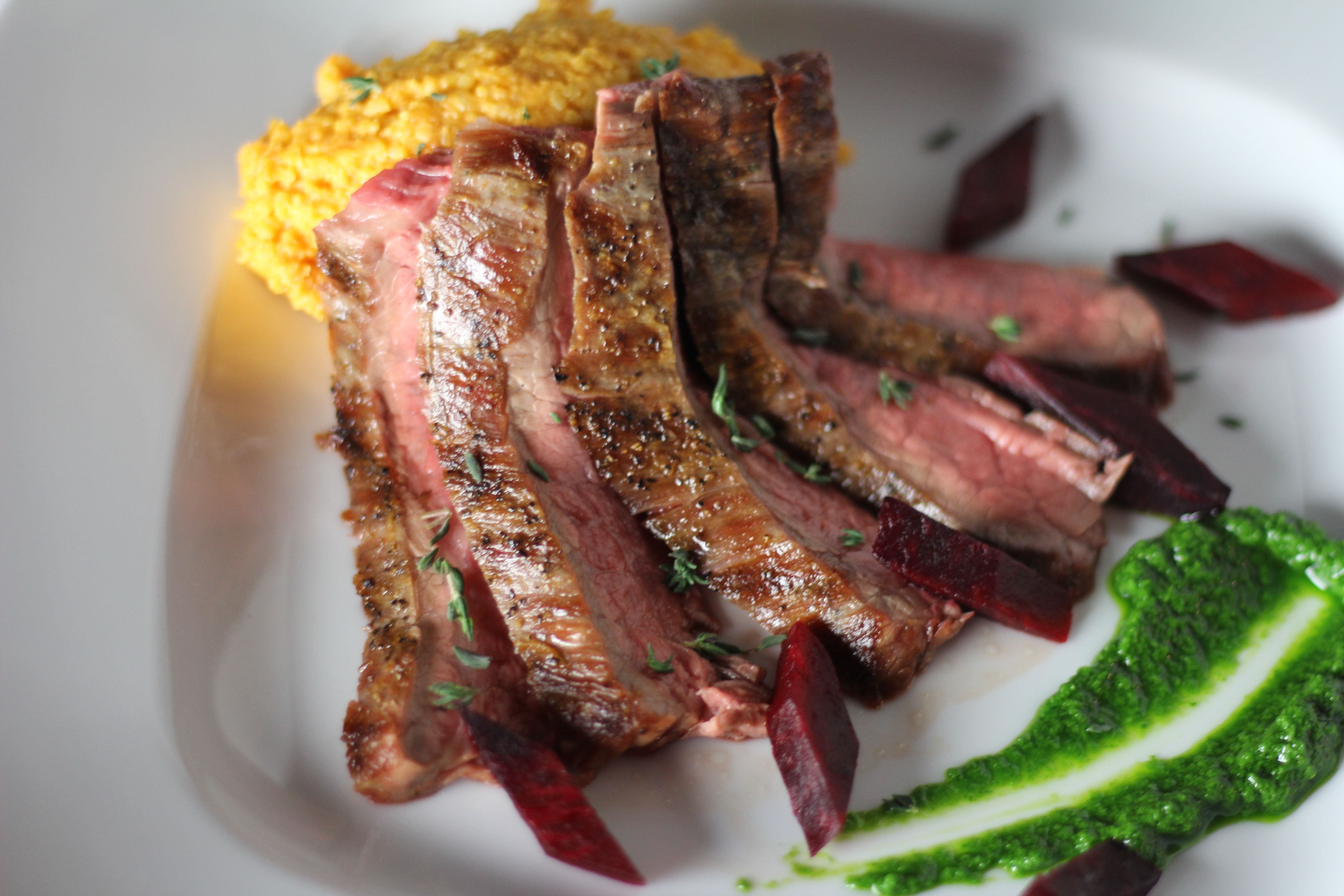 Gourmet sliced medium-rare steak garnished with thyme, roasted beet wedges, creamy orange squash mash and bright green herb puree on a white plate
