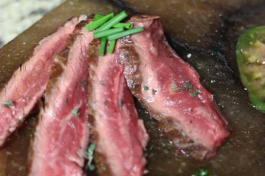 Close-up of juicy sliced medium-rare steak fanned on a dark board, topped with chopped chives and scattered herbs, with steak juices and a green tomato slice visible