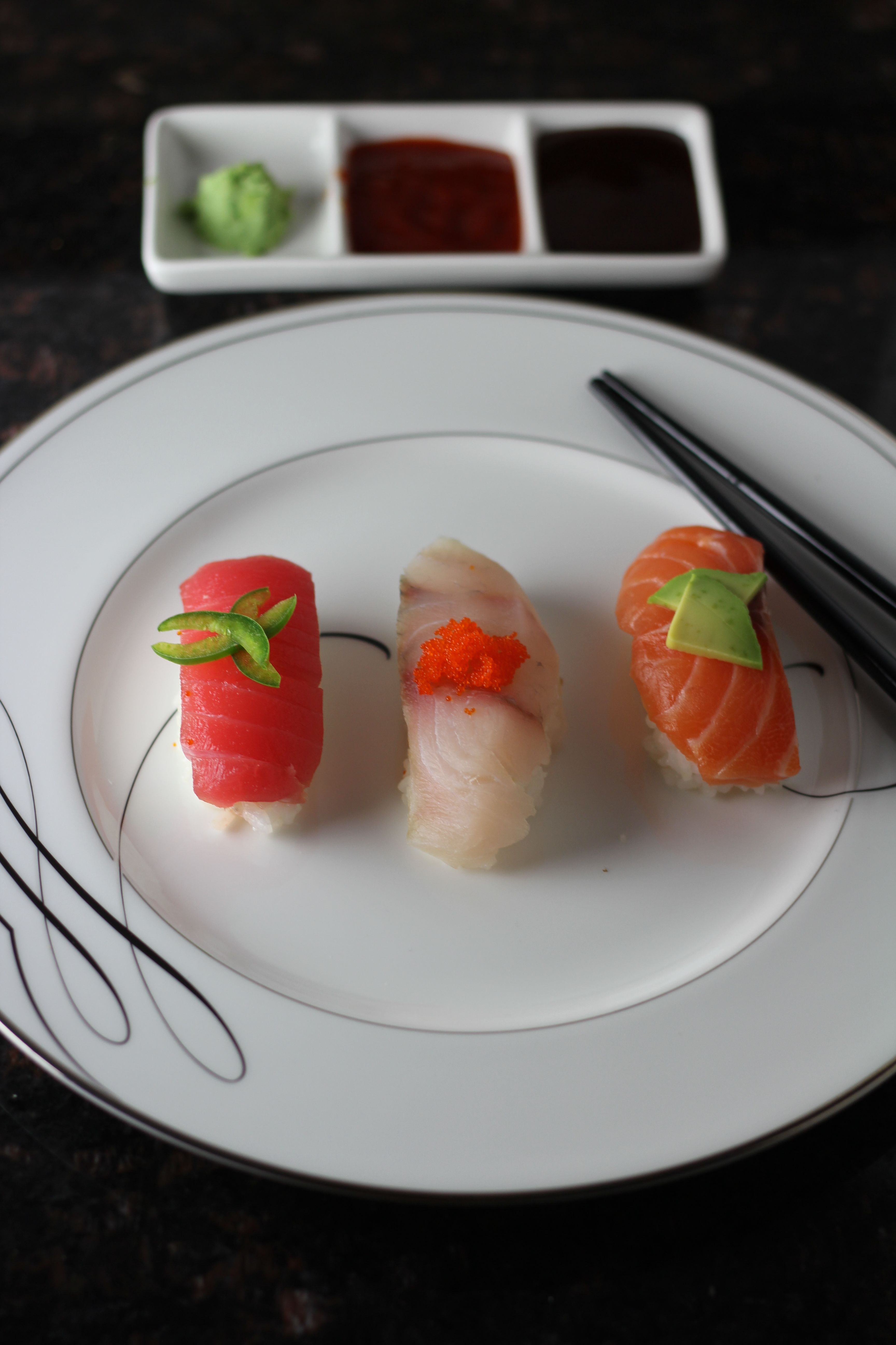 Three colorful nigiri sushi (tuna, white fish topped with orange tobiko, salmon with avocado) on a white plate with chopsticks and a three-compartment sauce dish with wasabi — Japanese sushi plate.