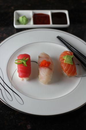 Three colorful nigiri sushi (tuna, white fish topped with orange tobiko, salmon with avocado) on a white plate with chopsticks and a three-compartment sauce dish with wasabi — Japanese sushi plate.