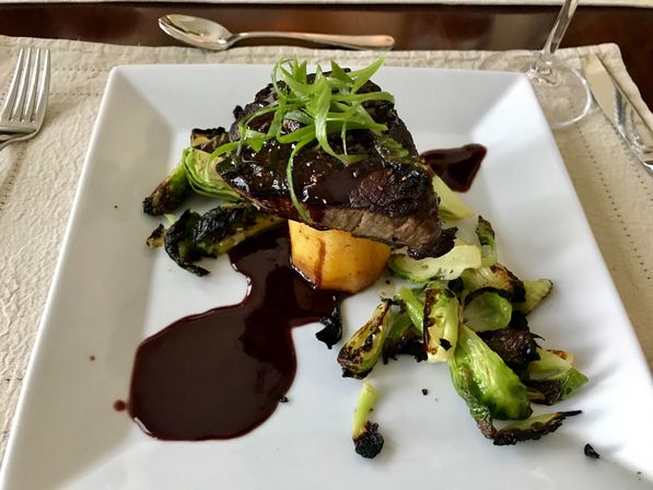 Seared steak atop a polenta cake with glossy red wine reduction, charred Brussels sprouts and scallion garnish on a white square plate at a table setting.