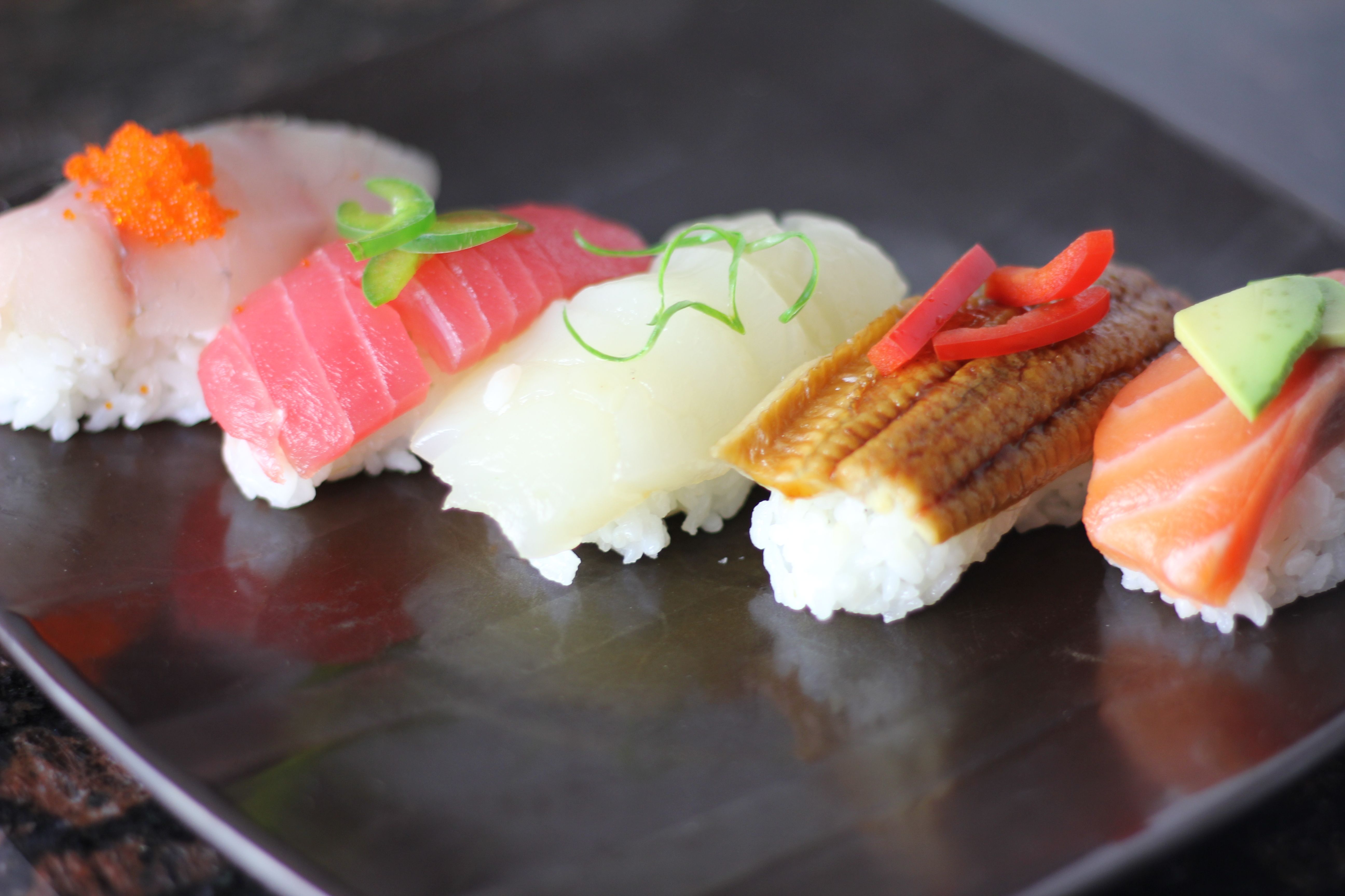 Colorful assorted nigiri sushi (tuna, scallop, eel, salmon and white fish) topped with roe and garnishes, arranged on a dark Japanese-style plate.