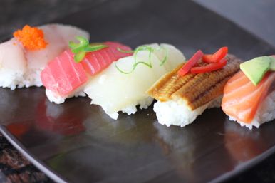 Colorful assorted nigiri sushi (tuna, scallop, eel, salmon and white fish) topped with roe and garnishes, arranged on a dark Japanese-style plate.