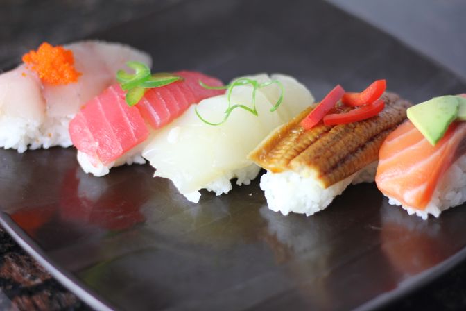 Colorful assorted nigiri sushi (tuna, scallop, eel, salmon and white fish) topped with roe and garnishes, arranged on a dark Japanese-style plate.