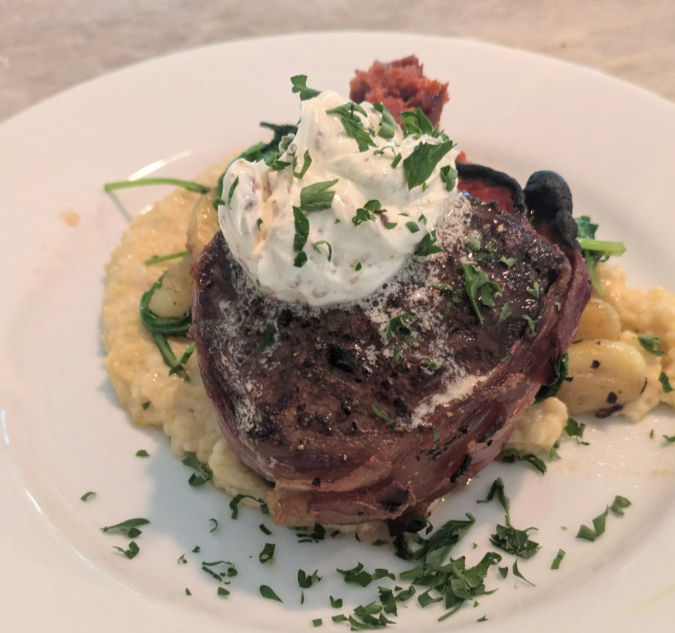 Prosciutto-wrapped filet mignon topped with herb cream served on creamy polenta with seared gnocchi and chopped parsley garnish