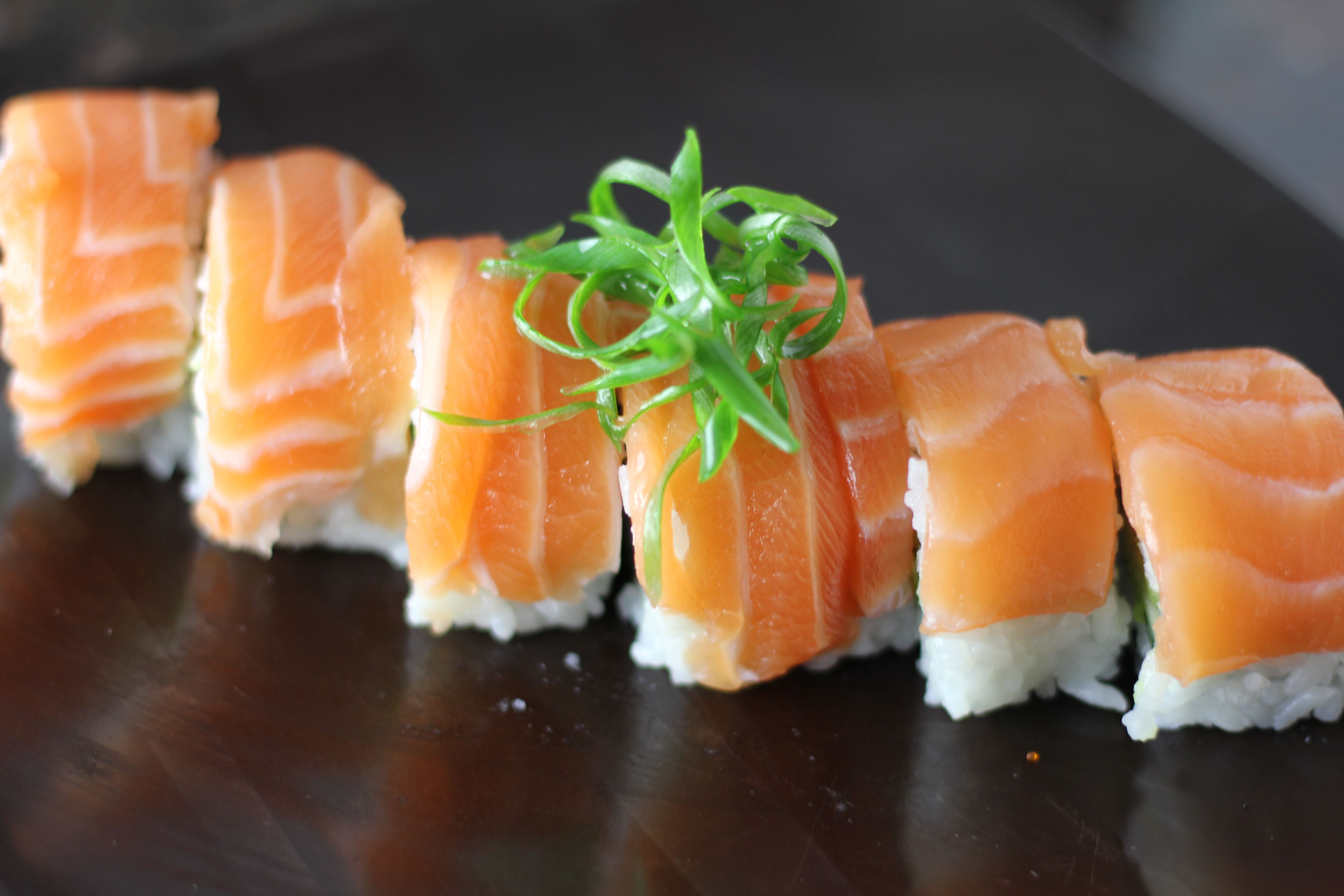 Fresh salmon-topped sushi rolls lined up on a sleek black plate with white rice and bright green scallion garnish