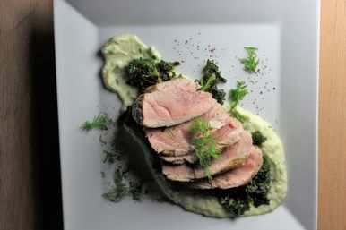 Gourmet sliced medium-rare pork tenderloin atop green pea purée and sautéed kale, garnished with fresh dill and cracked pepper on a white square plate