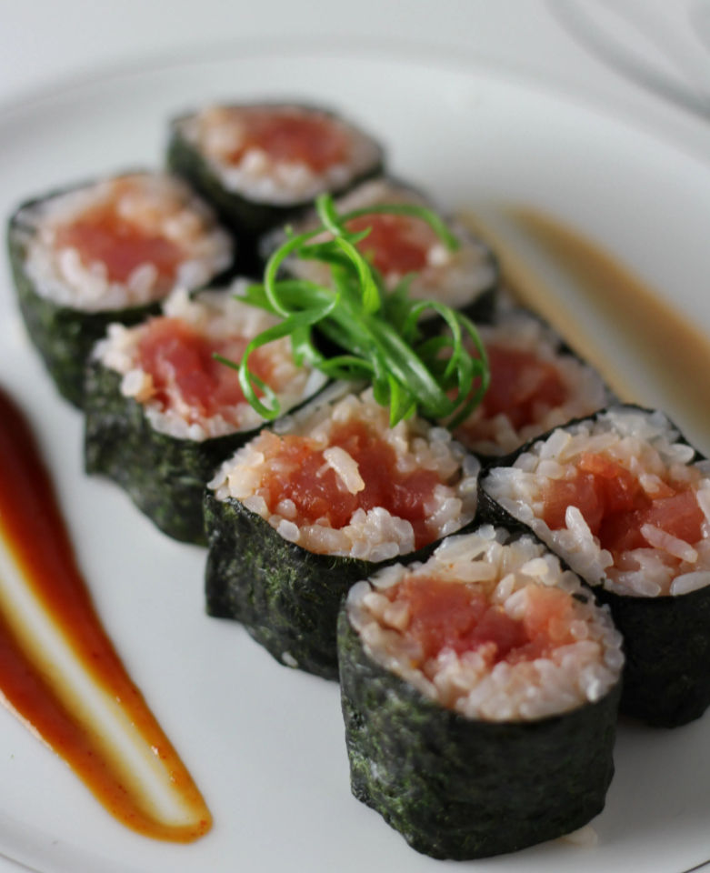 Close-up of tuna maki sushi rolls wrapped in nori on a white plate, garnished with curled scallions and two swooshes of soy-based sauce — Japanese-style sushi appetizer.