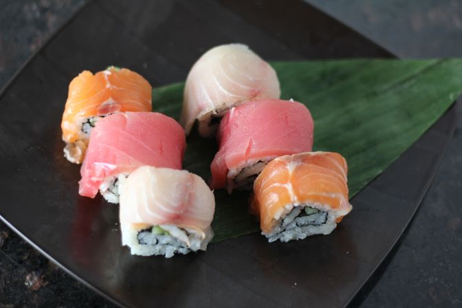 Six colorful Japanese sushi rolls topped with salmon, tuna, and white fish, arranged on a green leaf on a black plate