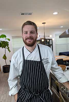 Smiling chef in a white coat and black-and-white striped apron leaning on a stainless stove in a bright modern open-plan kitchen with pendant lights and a potted fiddle-leaf fig