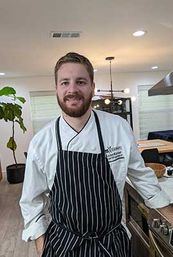Smiling chef in a white coat and black-and-white striped apron leaning on a stainless stove in a bright modern open-plan kitchen with pendant lights and a potted fiddle-leaf fig