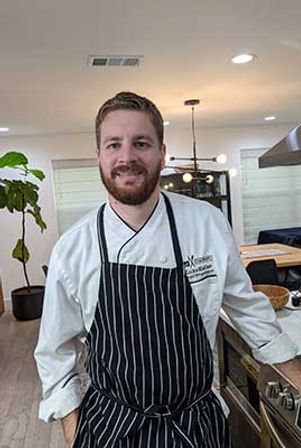 Smiling chef in a white coat and black-and-white striped apron leaning on a stainless stove in a bright modern open-plan kitchen with pendant lights and a potted fiddle-leaf fig