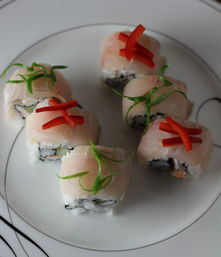 Six sushi rolls on a white plate topped with thin pale fish slices, red pepper strips and curled green scallion garnishes, with rice and seaweed visible in the centers.
