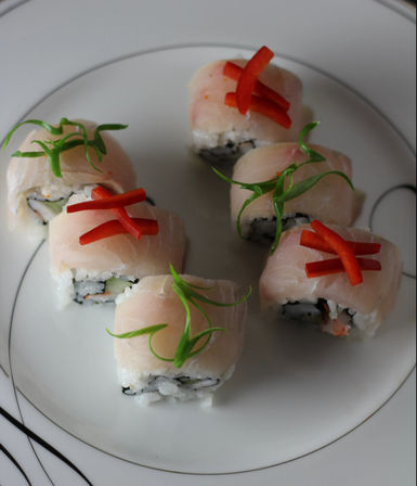 Six sushi rolls on a white plate topped with thin pale fish slices, red pepper strips and curled green scallion garnishes, with rice and seaweed visible in the centers.