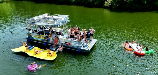Aerial view of people enjoying a sunny day on a two-level pontoon boat anchored on a green lake, with friends lounging on a large yellow floating mat, colorful inner tubes and small inflatable rafts nearby.
