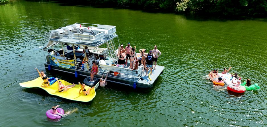 Aerial view of people enjoying a sunny day on a two-level pontoon boat anchored on a green lake, with friends lounging on a large yellow floating mat, colorful inner tubes and small inflatable rafts nearby.