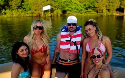 Group of five adults in swimsuits smiling on a boat at a sunny lake; man wears an American-flag life jacket and white cap, women in colorful bikinis and sunglasses with tree-lined shoreline in the background.
