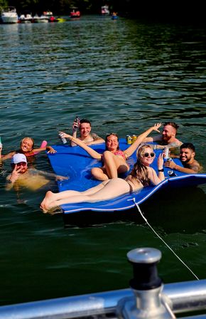 Group of friends lounging on a bright blue inflatable mat in a lake, sunbathing, smiling and holding canned drinks with boats and tree-lined shoreline in the background — summer boating and lake party scene.