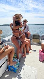 Friends on a pontoon boat at a sunny lake — two people in bikinis and straw hats hugging with drinks, another seated nearby, cooler and pack of bottled water on the deck.