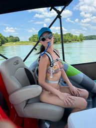 Young woman in a floral bikini and blue cap wearing heart-shaped sunglasses, sipping from a hydration tube while seated on a pontoon boat on a sunny lake with green shoreline.