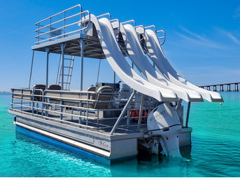 Pontoon party boat with four white water slides and outboard engine floating on clear turquoise water under a bright blue sky, bridge on the horizon.