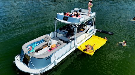 Aerial view of a sun-soaked pontoon boat anchored on a lake, people lounging on deck and a bright yellow floating mat