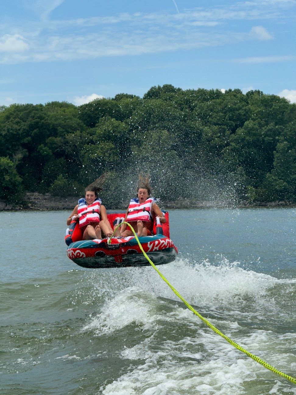 Two riders on a red inflatable towable tube soar over a boat wake on a sunny lake, water spraying around them and a wooded shoreline in the background.