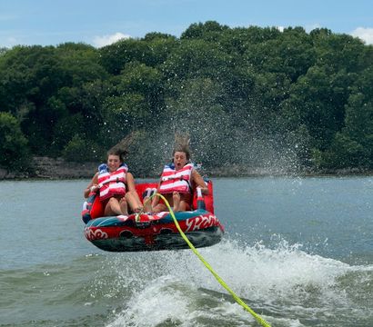 Two riders on a red inflatable towable tube soar over a boat wake on a sunny lake, water spraying around them and a wooded shoreline in the background.