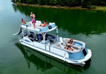 Aerial shot of a double-decker pontoon boat with a waterslide and people lounging on a calm green lake beside a tree-lined shoreline.