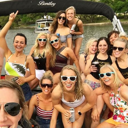 Group of smiling women on a pontoon boat at a lake, wearing swimsuits and sunglasses and holding canned drinks for a fun summer boat party selfie.