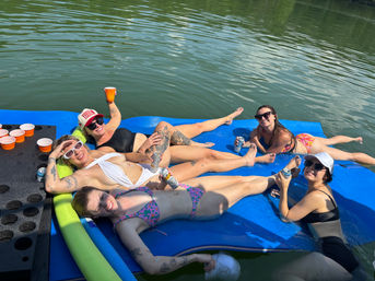 Five friends in swimsuits lounging on a blue floating mat in a calm lake, holding cans and cups, wearing sunglasses with orange cups in a drink holder nearby on a sunny summer day.