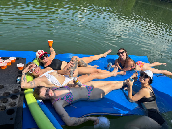 Five friends in swimsuits lounging on a blue floating mat in a calm lake, holding cans and cups, wearing sunglasses with orange cups in a drink holder nearby on a sunny summer day.