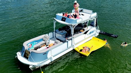 Aerial drone view of a pontoon boat anchored on a green lake, people sunbathing on the upper deck and a bright yellow floating mat, swimmers nearby in a summer boating scene