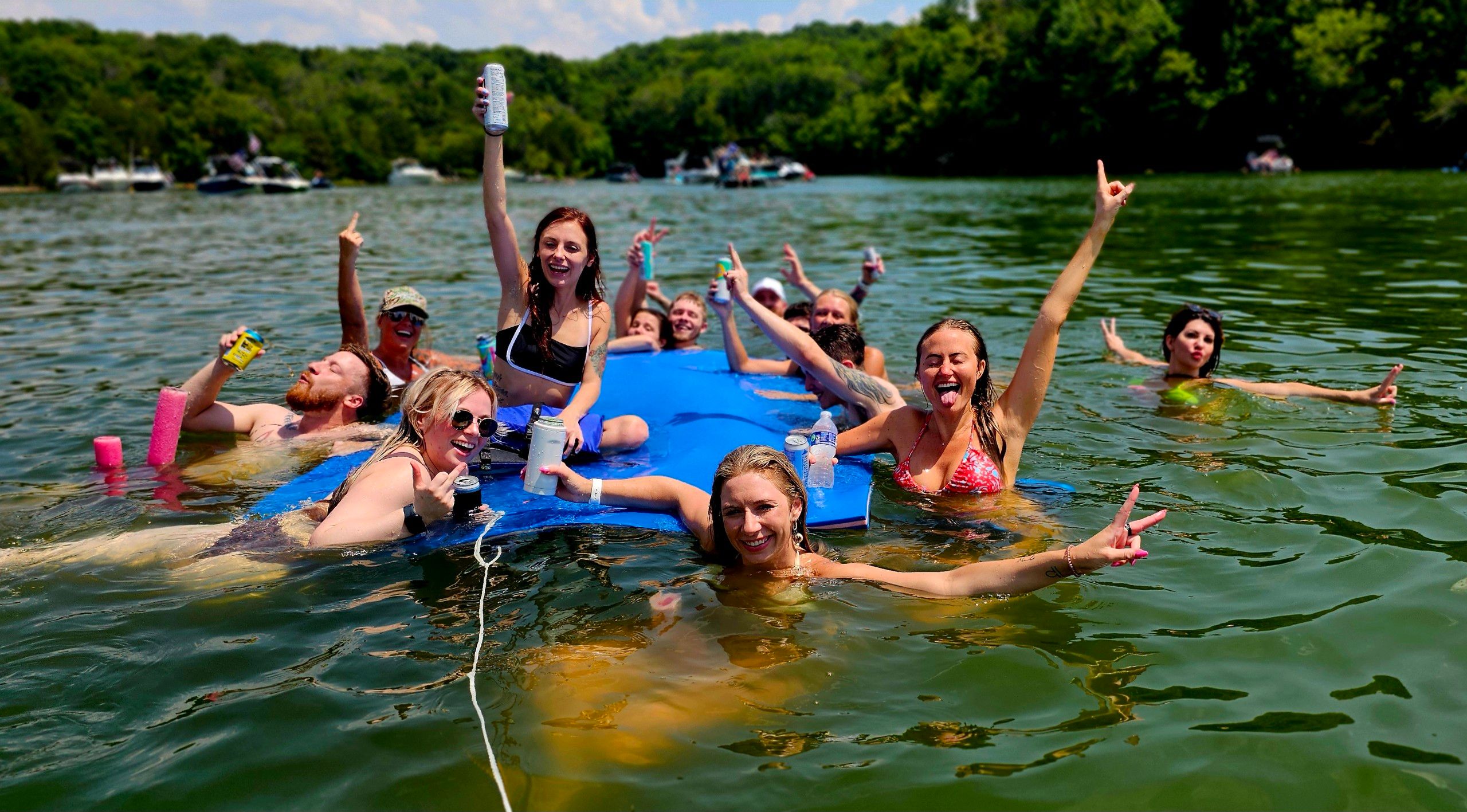 Group of friends enjoying a summer lake party on a large blue float, smiling and holding drinks in the water with boats and a tree-lined shoreline in the background.
