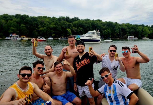 Smiling group of friends on a pontoon boat at a summer lake party, raising beer cans with other boats and a tree-lined shoreline in the background.