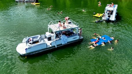Aerial view of a pontoon boat anchored on a green lake with people sunbathing on the roof, swimmers lounging on a blue floating mat and others splashing nearby on a sunny day.