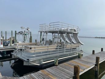 Two-level pontoon boat with upper sun deck and twin water slides docked at a wooden pier on calm coastal waters under a hazy sky.