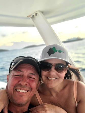 Boat selfie of two smiling adults wearing caps and sunglasses on the ocean at sunset with island silhouettes in the background.